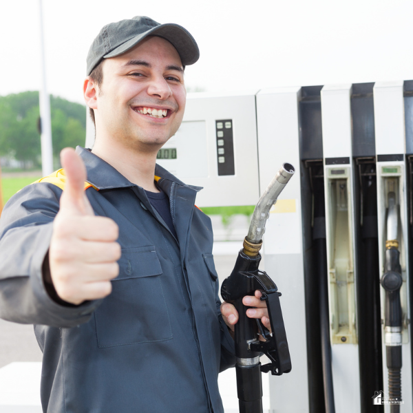 Smiling gas station worker in uniform gives a thumbs up while holding a fuel nozzle.
