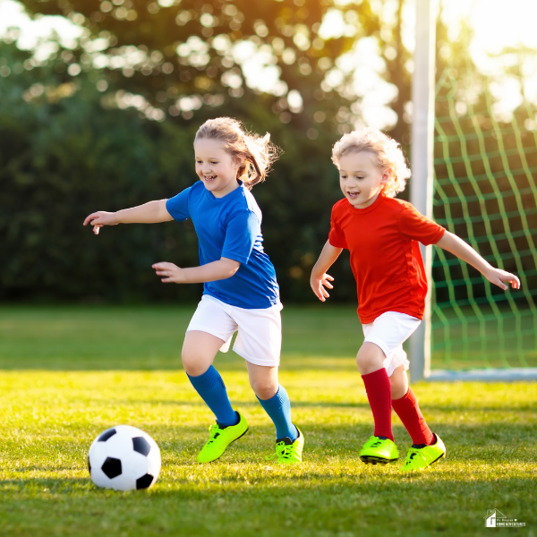 Two young children in colorful uniforms joyfully run after a soccer ball on a grassy field.