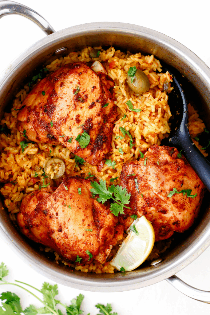Overhead view of a full pot of arroz con pollo with three pieces of chicken, rice, olives, cilantro, and a serving spoon ready to serve.