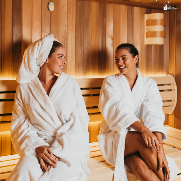 Two women wearing white robes sit and smile while relaxing inside a wood-paneled sauna.