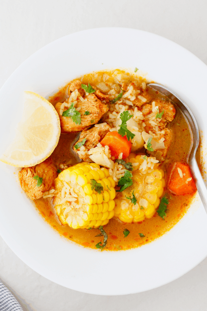 Detailed close-up of Puerto Rican chicken soup, highlighting chunks of chicken, corn, carrots, potatoes, and rice in a golden broth.
