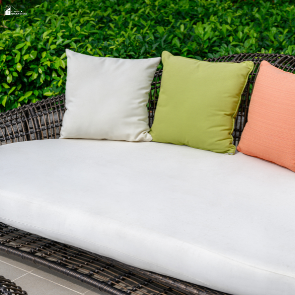 Close-up of a wicker patio sofa with white cushions and three colorful throw pillows set against a green hedge.
