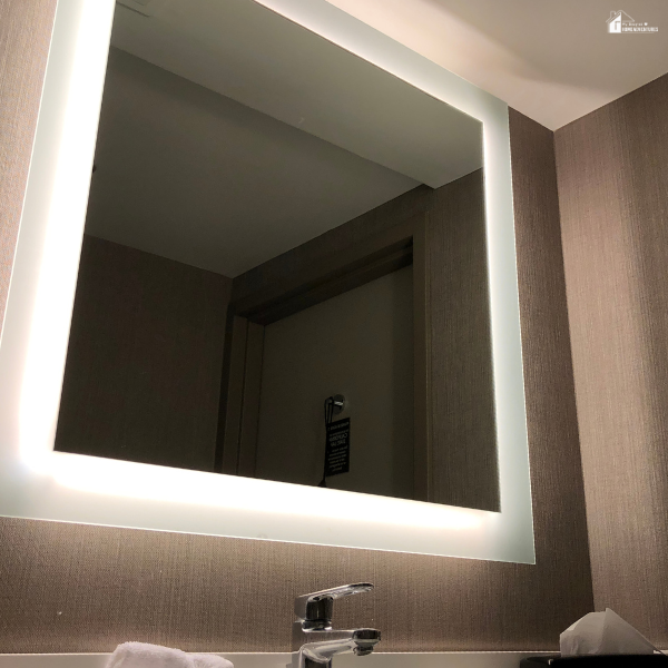 A square LED-backlit mirror mounted above a bathroom sink with a beige wall.