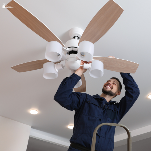 A man stands on a ladder and carefully deep cleans the blades of a modern ceiling fan in a well-lit room.