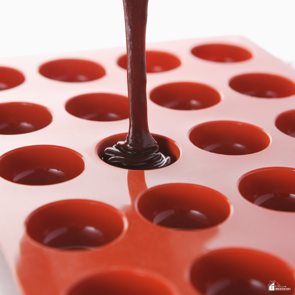A close-up of melted chocolate being poured into a red silicone mold with round cavities.