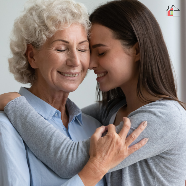 A young woman gently embracing her elderly mother, both smiling with closed eyes, sharing a warm and affectionate moment in a bright home setting.