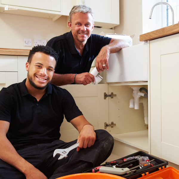 Two plumbers smiling while repairing plumbing beneath a kitchen sink with tools.
