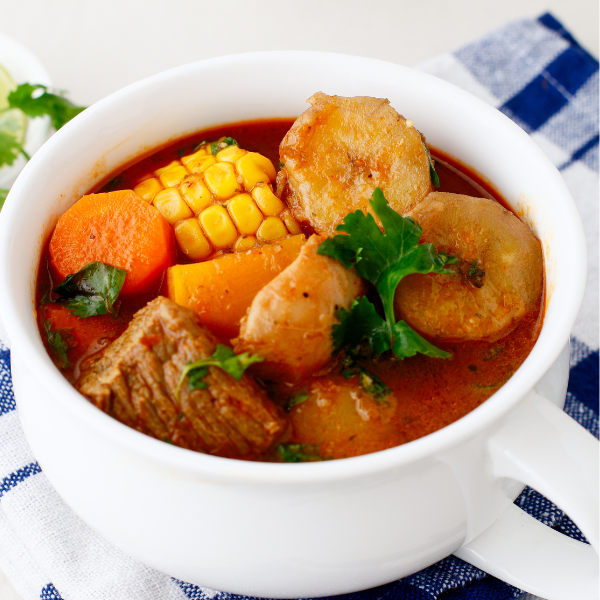 A white bowl filled with Puerto Rican beef sancocho featuring tender chunks of beef, corn, carrots, and potatoes in a rich, red broth garnished with fresh cilantro.