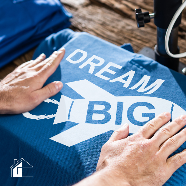 Hands pressing a blue T-shirt with a white “Dream Big” rocket graphic design on it during a heat-transfer printing process.