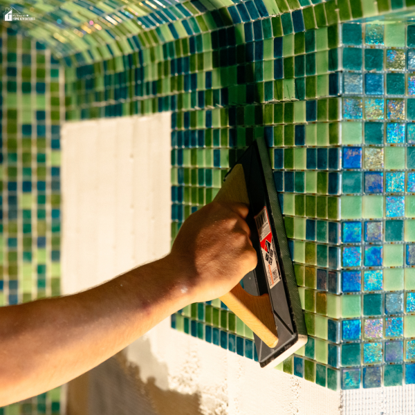 Person applying grout to a wall of green and blue glass mosaic tiles using a float tool.