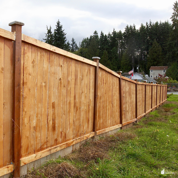 A long wooden privacy fence installed on a grassy slope with trees and houses in the background, showing professional fencing work.