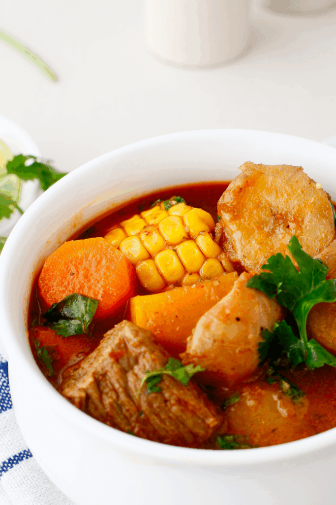 A white bowl filled with Puerto Rican beef sancocho featuring tender chunks of beef, corn, carrots, and potatoes in a rich, red broth garnished with fresh cilantro.