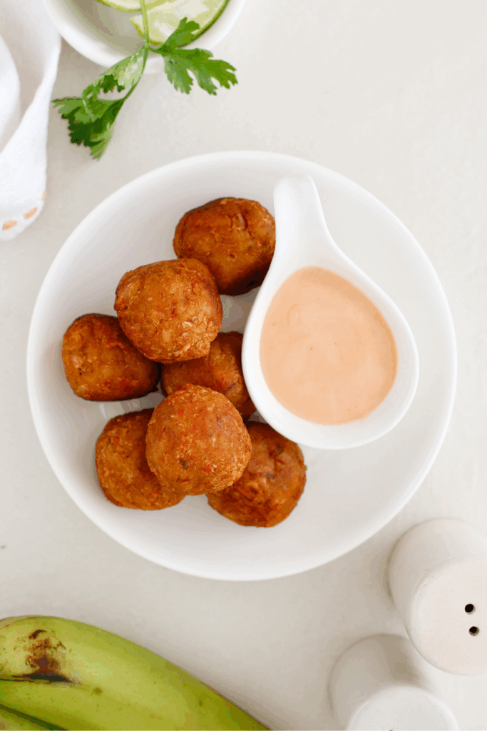 A white plate filled with crispy mofongo balls served with a side of creamy dipping sauce, garnished with fresh lime and herbs.