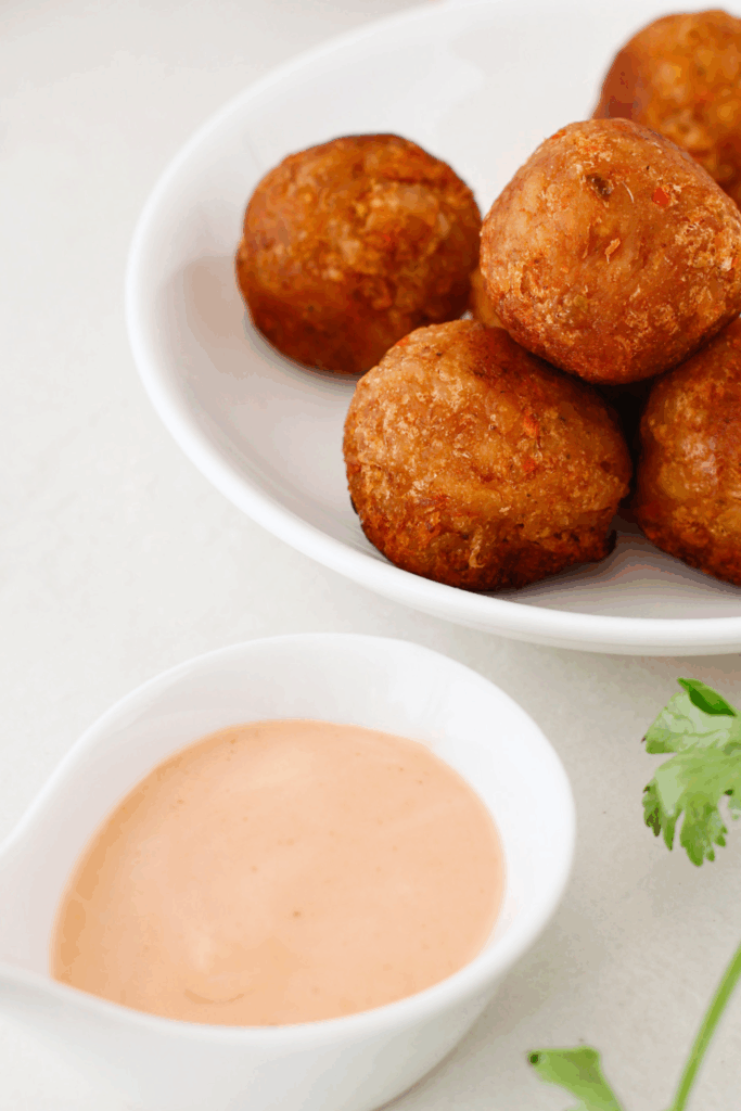 A bowl of freshly fried mofongo balls with a small dish of dipping sauce and a sprig of cilantro nearby for garnish.
