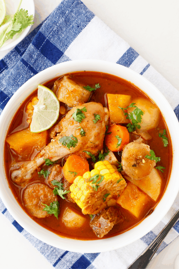 Top-down view of a bowl of Puerto Rican beef sancocho with chicken drumstick, vegetables, corn, and cilantro served with lime wedges on the side.