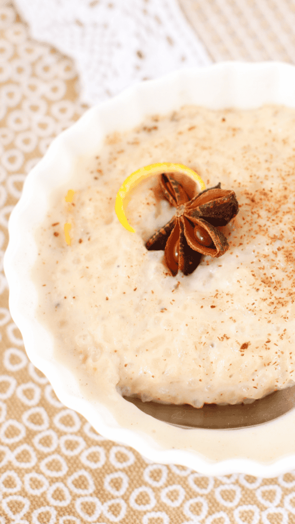Close-up of creamy arroz con leche topped with star anise, citrus peel, and cinnamon in a white scalloped bowl.