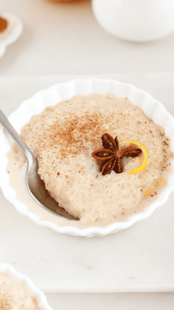 Creamy arroz con leche served in a white scalloped bowl with a spoon, topped with star anise, citrus peel, and a dusting of cinnamon.