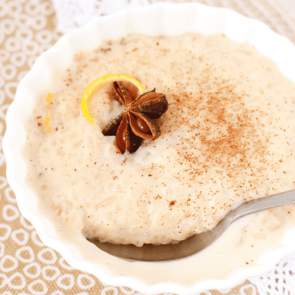 Close-up of creamy arroz con leche topped with star anise, citrus peel, and cinnamon in a white scalloped bowl.