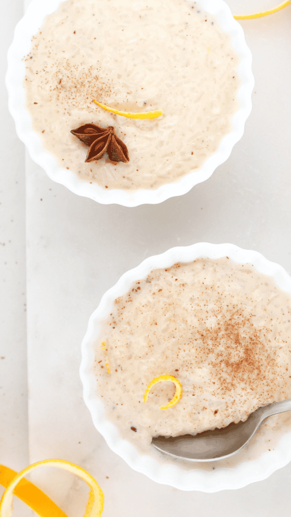 Two bowls of creamy arroz con leche garnished with cinnamon, star anise, and citrus peel, with a spoon resting in one bowl.