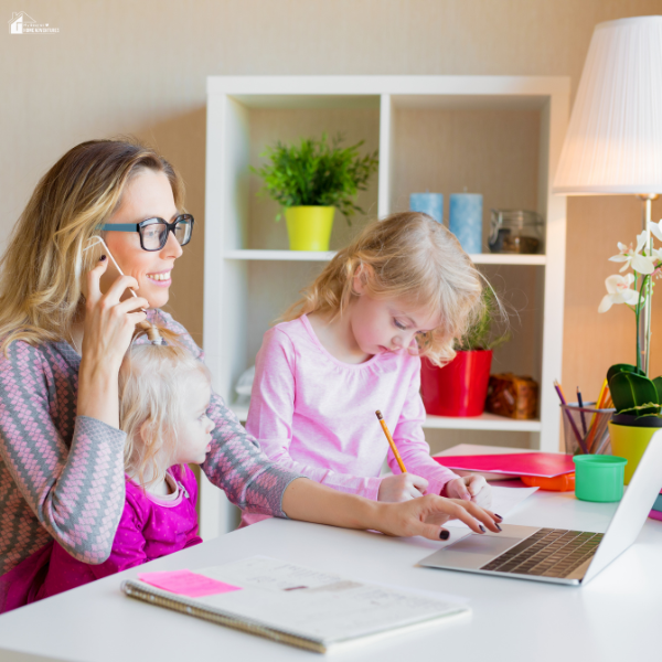 A mom works on a laptop while caring for her children, balancing parenting and running a business from home.