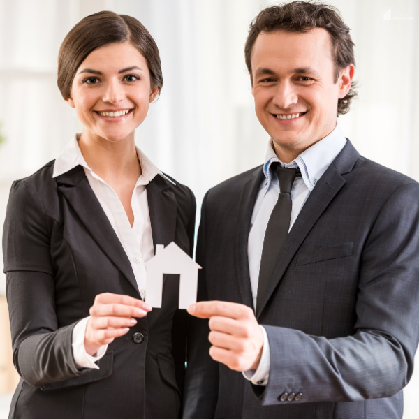 A professional man and woman in business attire smiling and holding a small paper house symbol, representing real estate professionals.