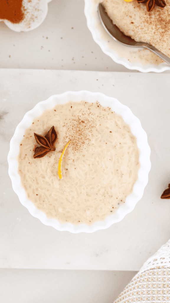 Overhead view of creamy arroz con leche in a white bowl, garnished with star anise, citrus peel, and a light sprinkle of cinnamon.