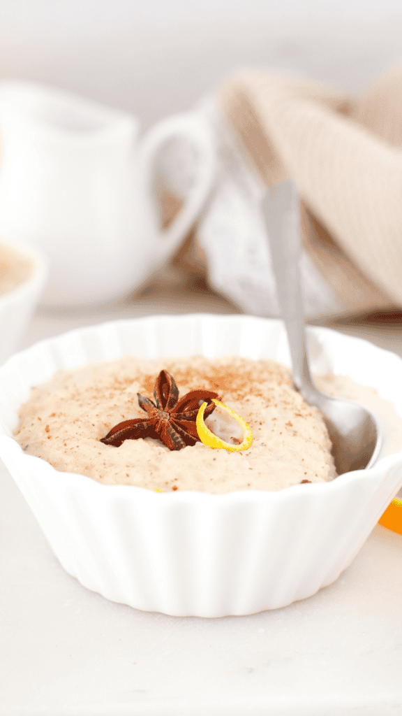 Creamy arroz con leche in a white scalloped bowl topped with star anise, citrus peel, and cinnamon, with a spoon resting along the side.