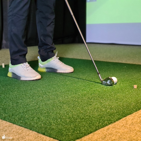 A golfer standing on a turf mat, preparing to hit a ball with a golf club indoors.