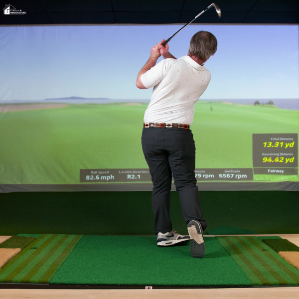A man mid-swing inside a golf simulator room with a virtual fairway projected on the screen.