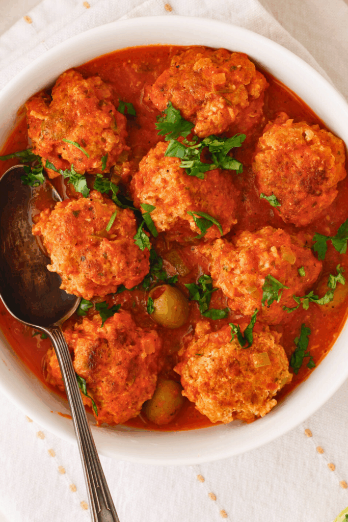 Puerto Rican meatballs simmered in a rich tomato sauce with sofrito, sazon, and green olives, also known as albondigas guisadas, garnished with fresh cilantro in a serving bowl