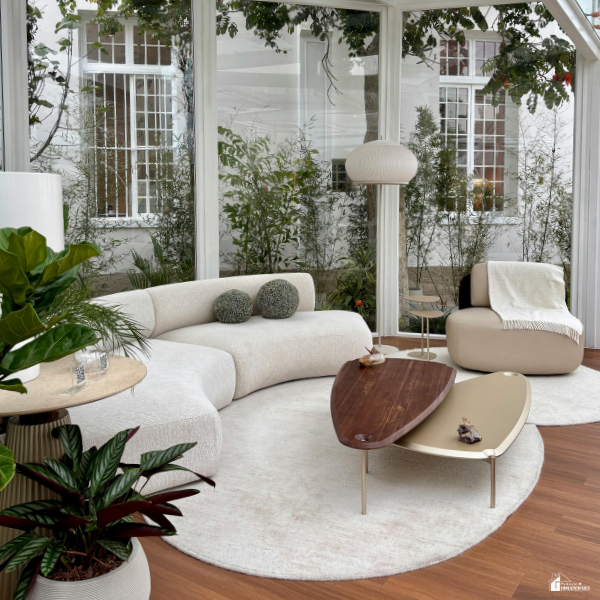 Modern glass-enclosed sunroom with curved sofa, indoor plants, and wood coffee tables creating a bright summer garden room retreat.