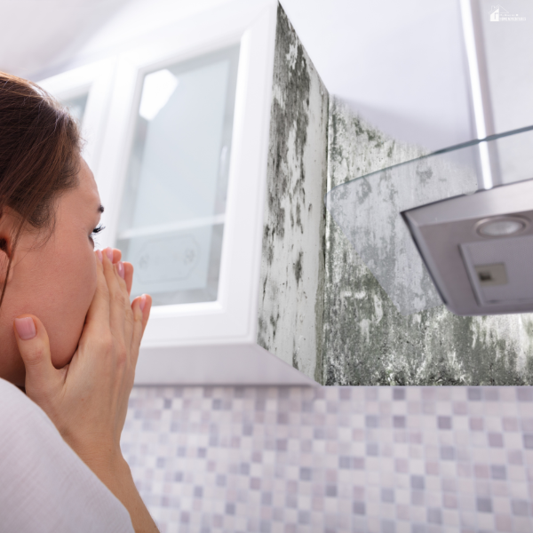 Woman covering her nose while looking at visible mold growth on a kitchen cabinet wall near a stove vent.