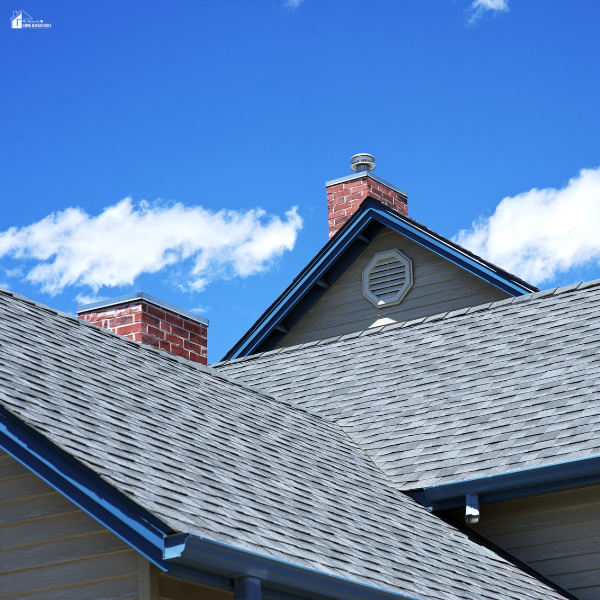 Close-up of a home with gray asphalt shingles and brick chimneys under a blue sky, highlighting roofing material and architectural style options.