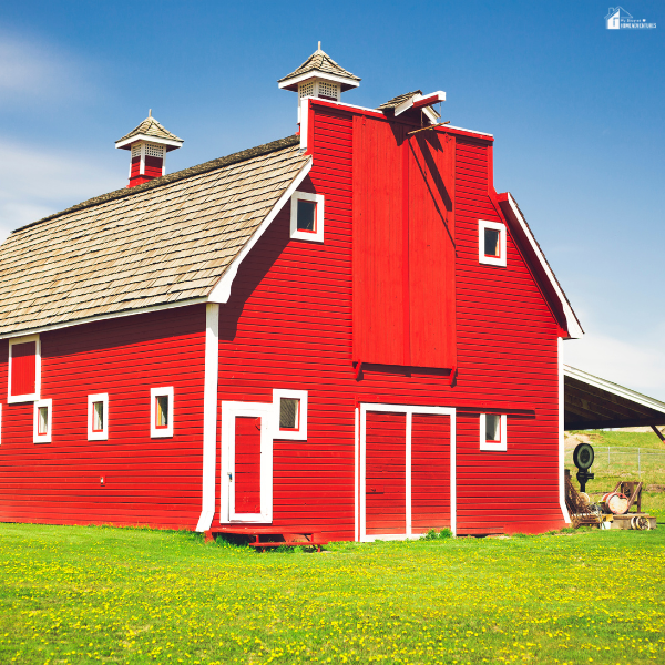 A bright red pole barn with white trim and cupolas sits on a green field under a clear blue sky.
