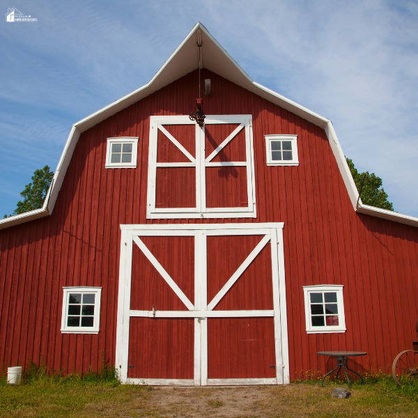 A classic red pole barn with white framed windows and large double doors stands beneath a sunny blue sky.
