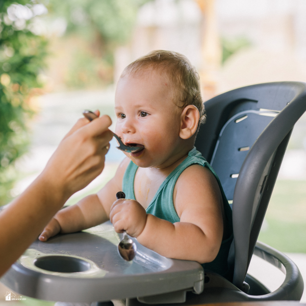 Baby sitting in a high chair being fed with a spoon by a caregiver during mealtime to support healthy first-year nutrition.