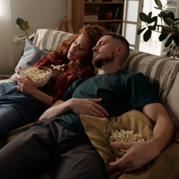 Tired couple asleep on a couch with snacks in their hands, illustrating exhaustion that can result from poor sleep routines.