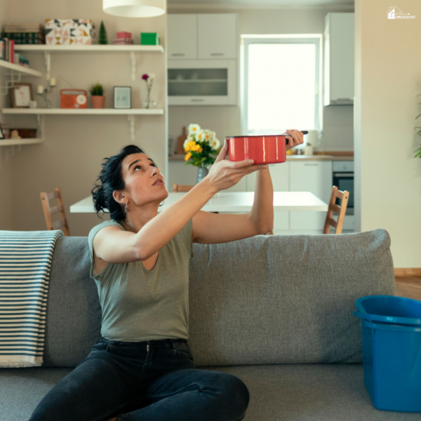 Person sitting on a couch holding a pot to catch a ceiling leak, showing basic home maintenance that prevents bigger problems.