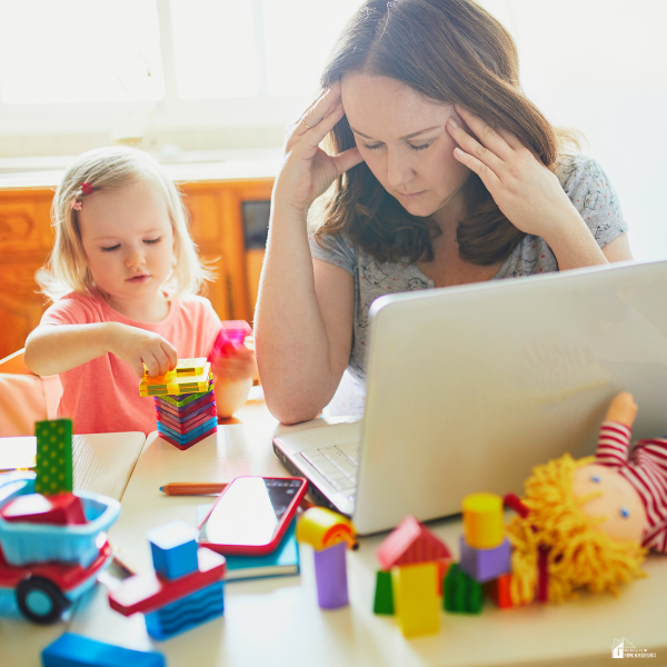 Parent working on a laptop while a child plays nearby, showing the unseen effort of balancing home responsibilities and daily life.