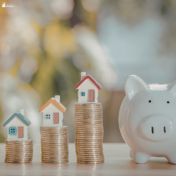 Small model houses stacked on coins next to a piggy bank, representing saving money for a home with irregular income.