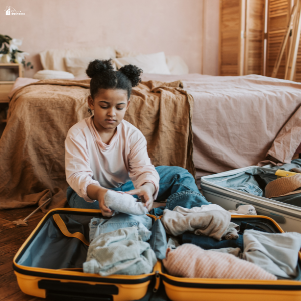Child organizing clothes into a suitcase on the floor while preparing for a long distance move on a budget with family.