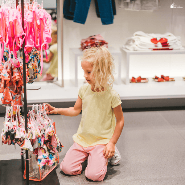Young child browsing children’s swimsuits in a clothing store during a family summer shopping trip on a budget.