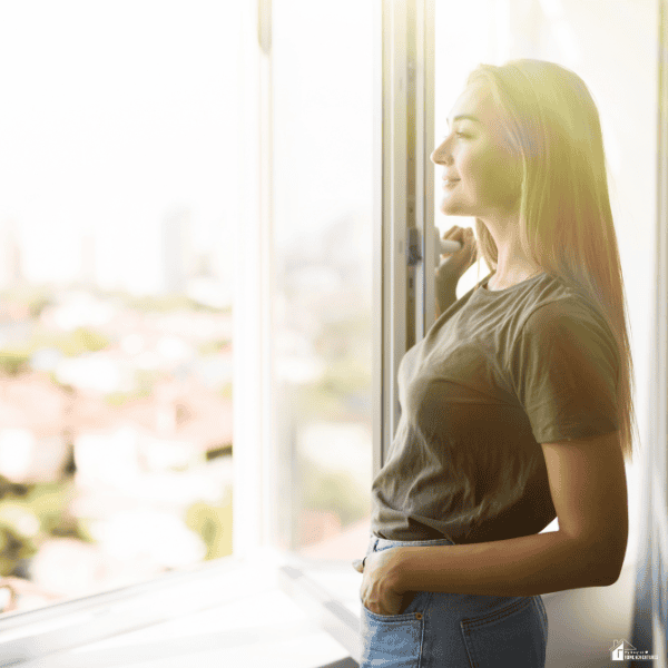 A woman stands beside an open window, taking in fresh air as part of a clean and healthy home environment.