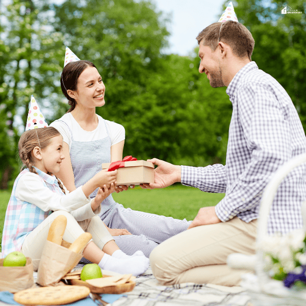 A father gives his young daughter a wrapped present during a family picnic, highlighting a thoughtful gift for daughters on a special occasion.