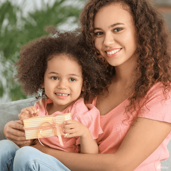 A young girl smiles while holding a wrapped present beside her mother, representing a heartfelt gift for daughters to celebrate special moments.
