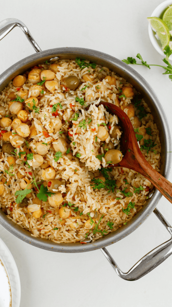 Overhead view of Puerto Rican arroz con garbanzos with chickpeas, green olives, fresh cilantro, and a wooden spoon in a large skillet.