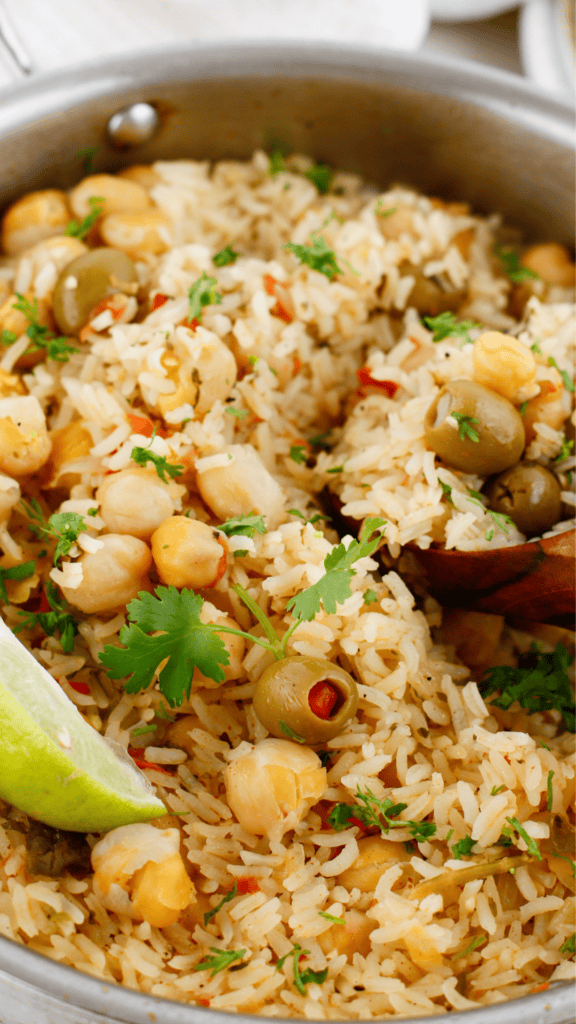 Close-up of fluffy Puerto Rican rice with chickpeas, green olives, fresh cilantro, and a lime wedge served in a skillet.