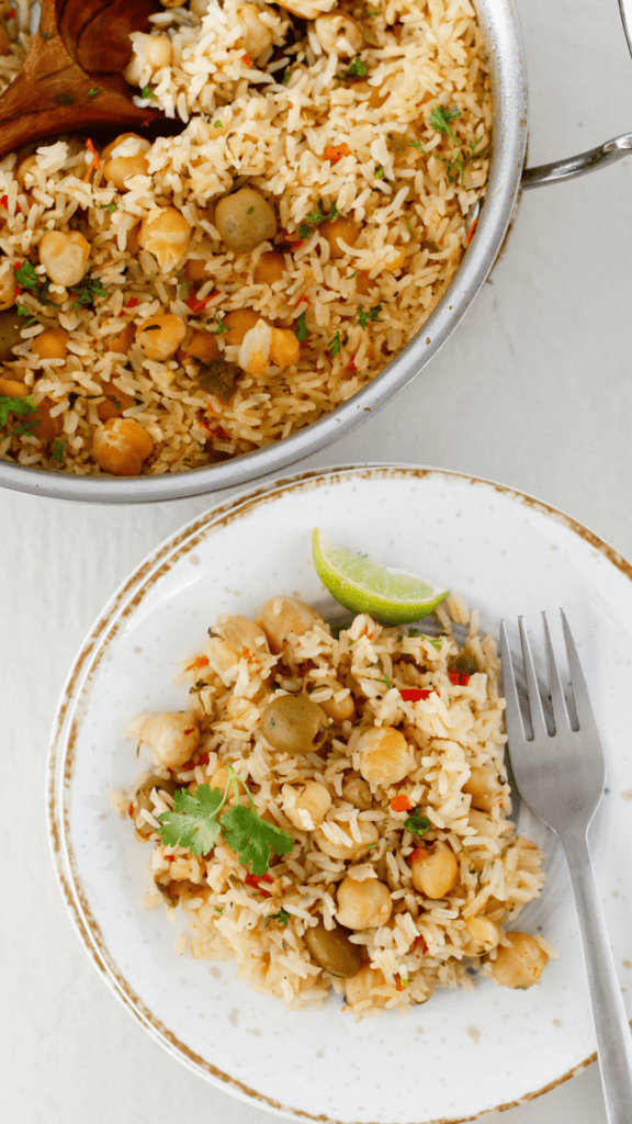 A plated serving of Puerto Rican arroz con garbanzos with chickpeas, green olives, cilantro, and a lime wedge beside a skillet of rice.