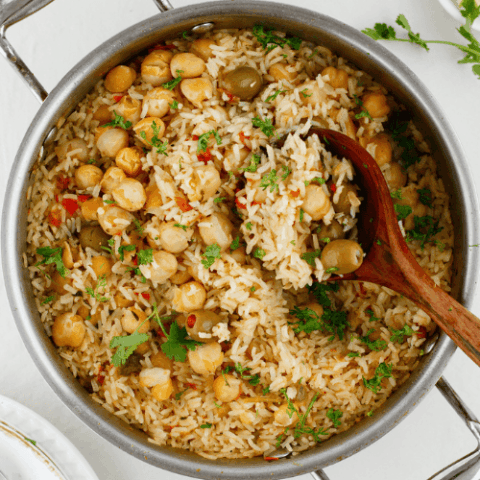 Overhead view of Puerto Rican arroz con garbanzos with chickpeas, green olives, fresh cilantro, and a wooden spoon in a large skillet.