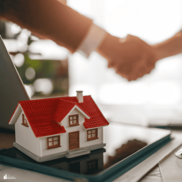 A buyer and seller complete a real estate closing with a handshake beside a model home representing the completed property sale.